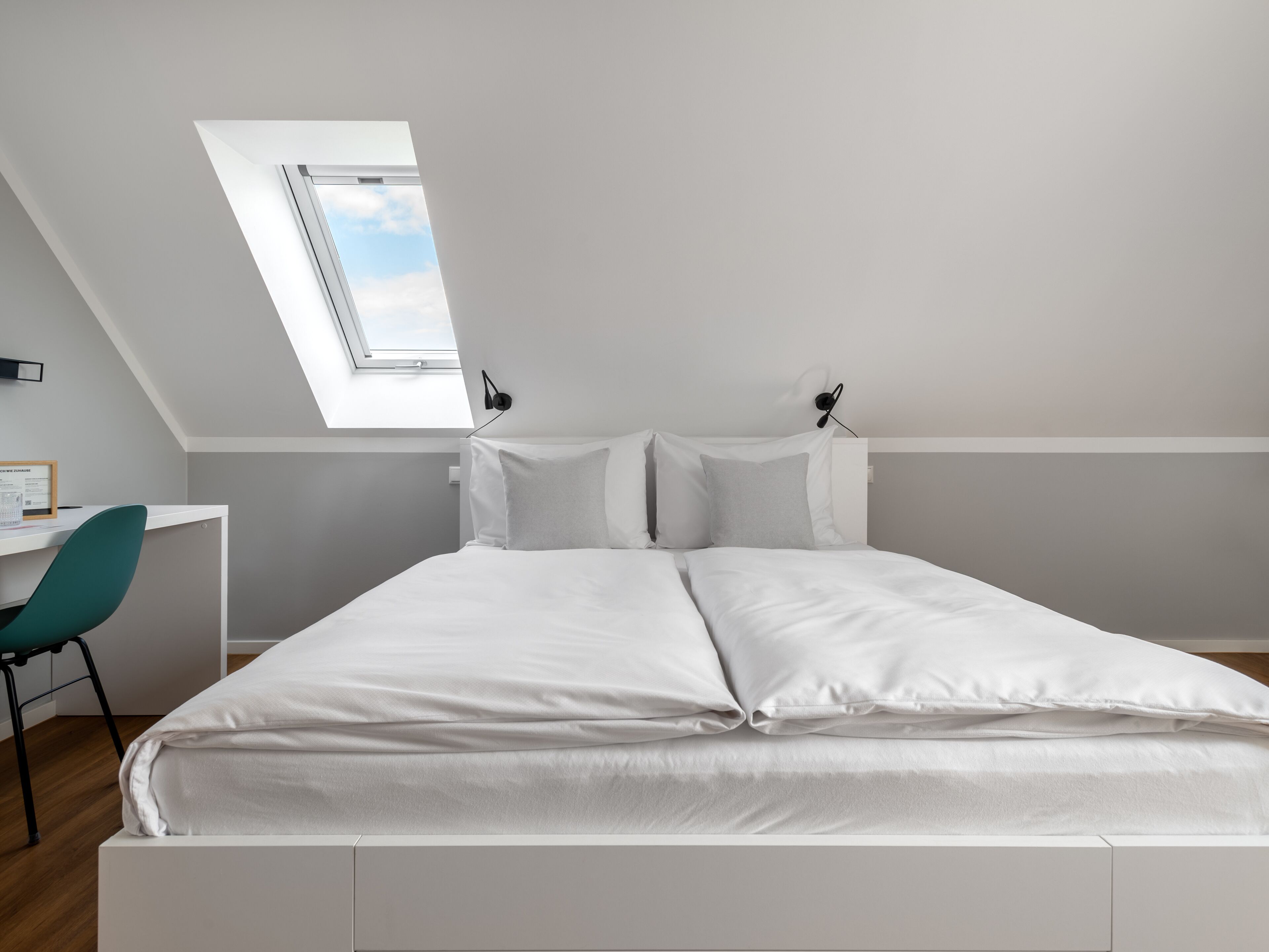 Ein modernes Schlafzimmer mit Dachfenster, einem Bett mit weißer Bettwäsche, einem Schreibtisch mit Stuhl und einer Wandlampe.