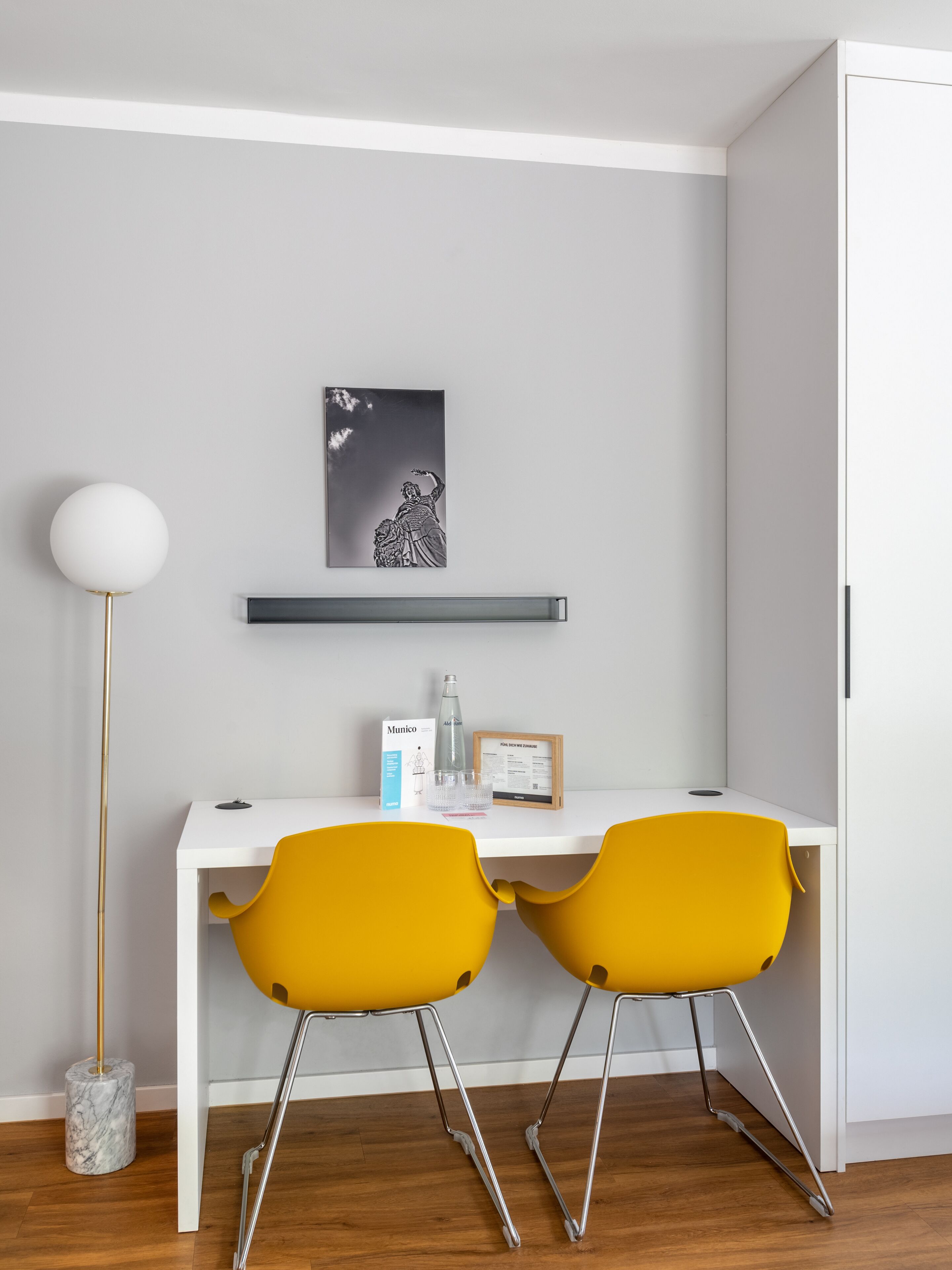 Una habitación moderna con un escritorio blanco, dos sillas amarillas, suelo de mármol, una lámpara de pie blanca y un cuadro blanco y negro en la pared.