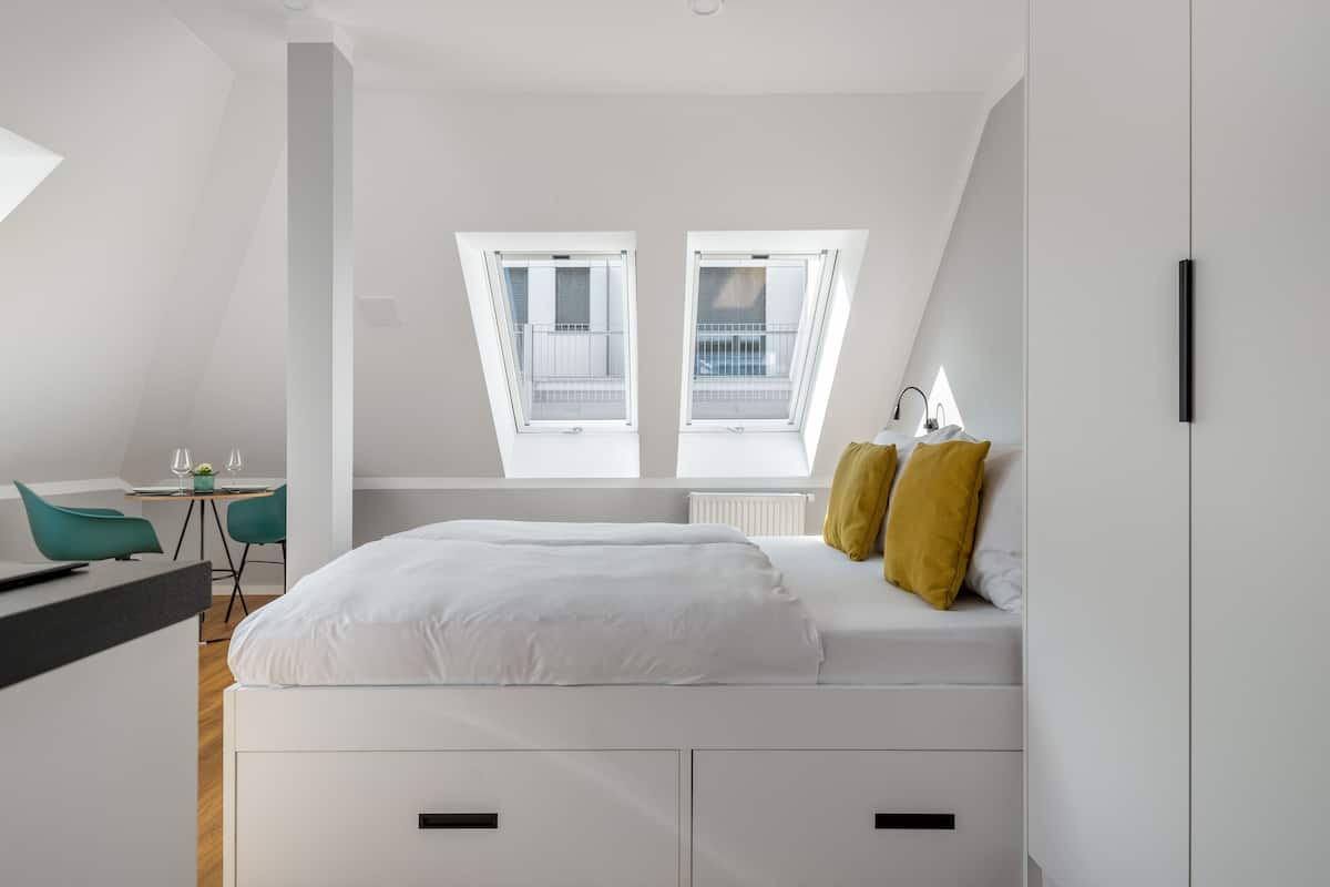 Une chambre à coucher moderne avec une lucarne, un lit recouvert d’une literie blanche, deux oreillers jaunes et un bureau avec une chaise.