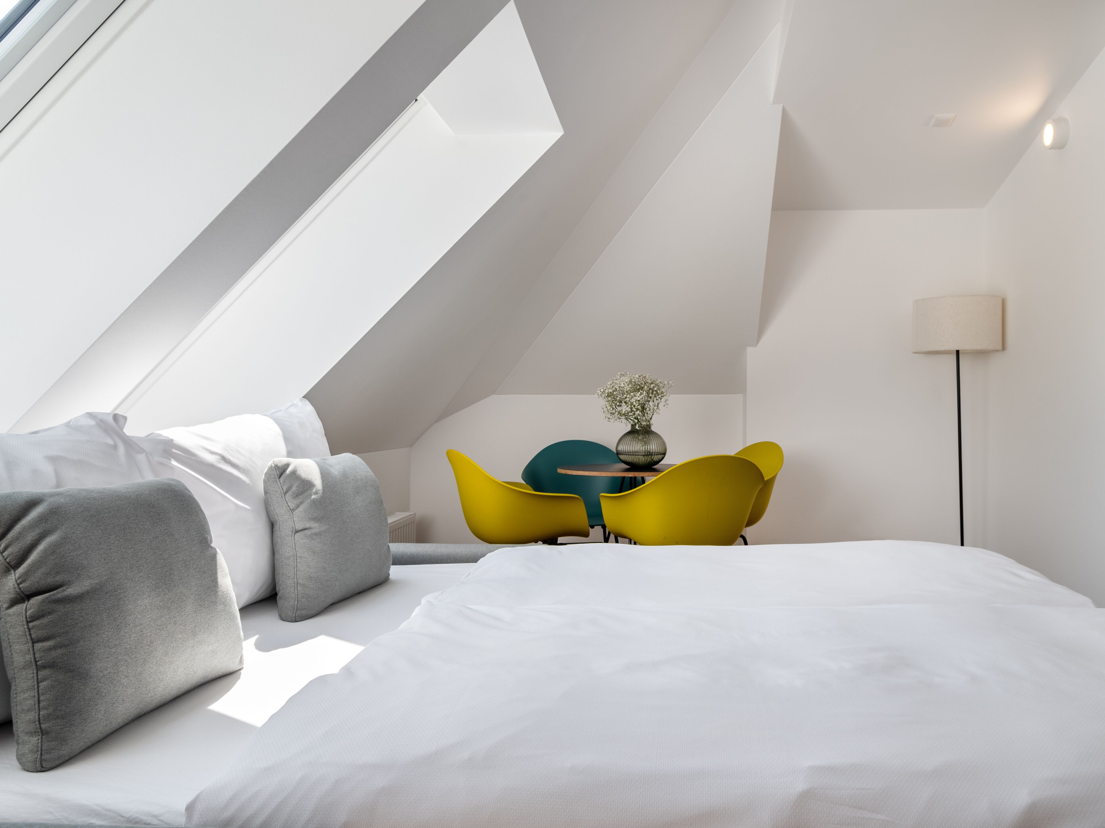 Une pièce moderne et bien éclairée, avec un plafond en pente, un lit recouvert de linge de maison blanc, une petite table ornée d’un vase et des chaises jaunes.