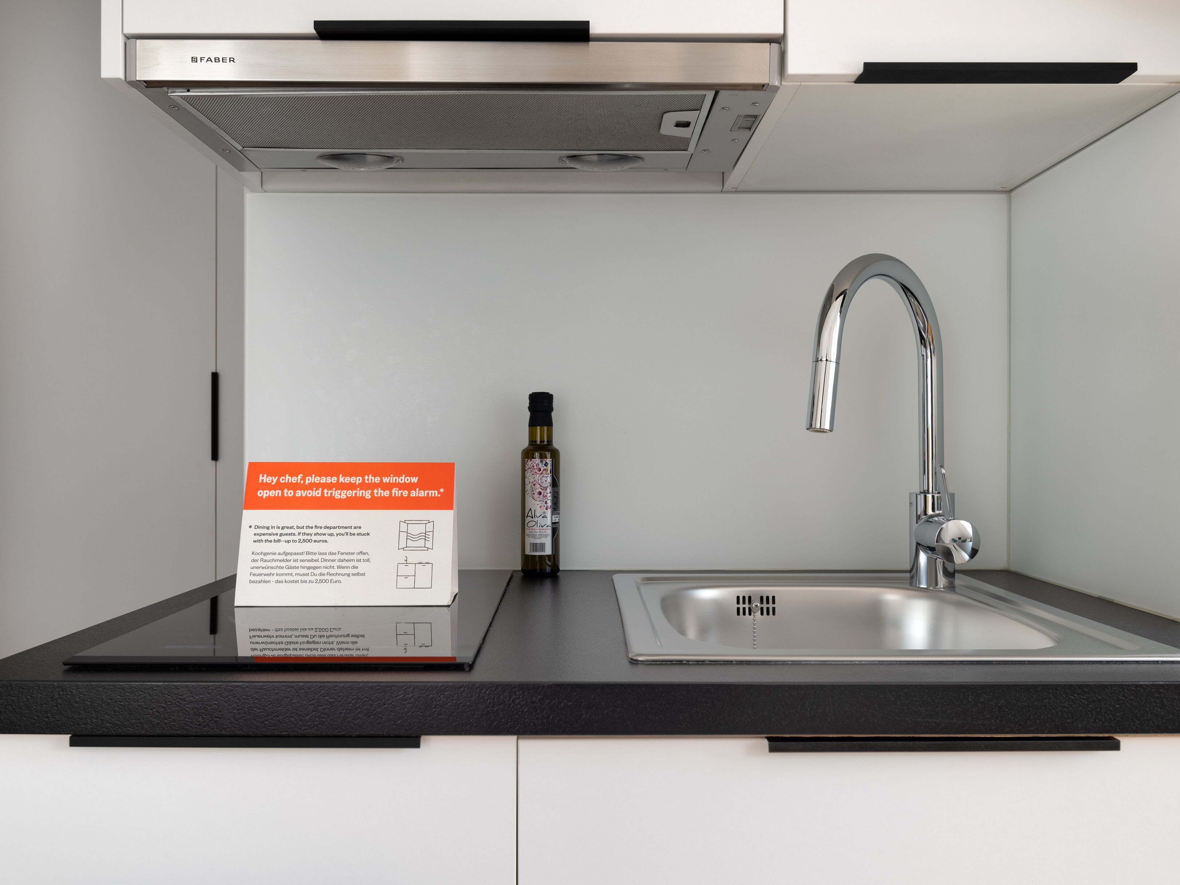 A modern kitchen with a stainless steel sink, a faucet, and a Faber range hood. There's a bottle of olive oil and a sign advising to keep the window open to avoid triggering the fire alarm.