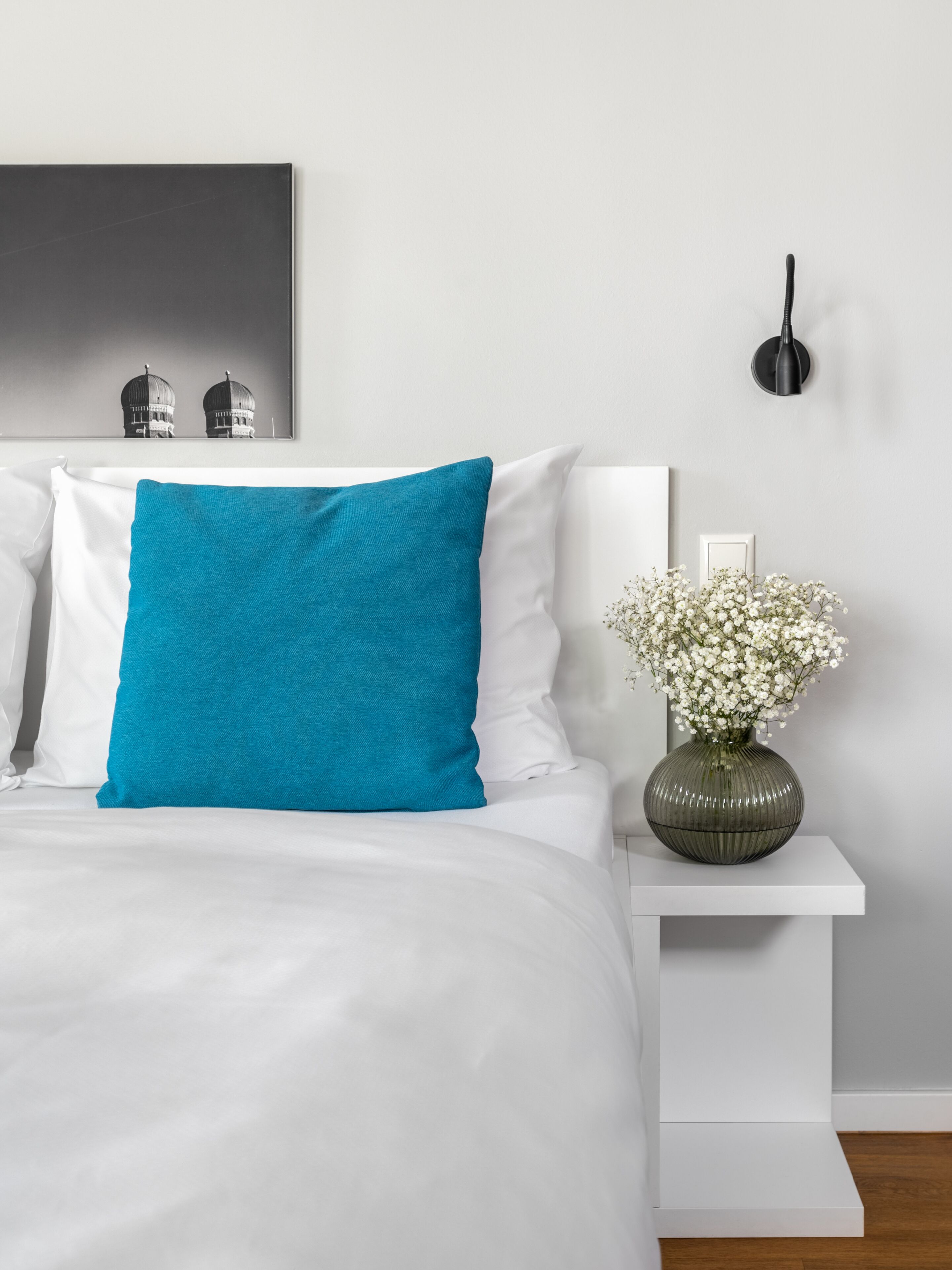 Una cama bien hecha con almohadas blancas y verde azulado, una mesita de noche blanca con un jarrón de flores blancas y una lámpara fijada a la pared.