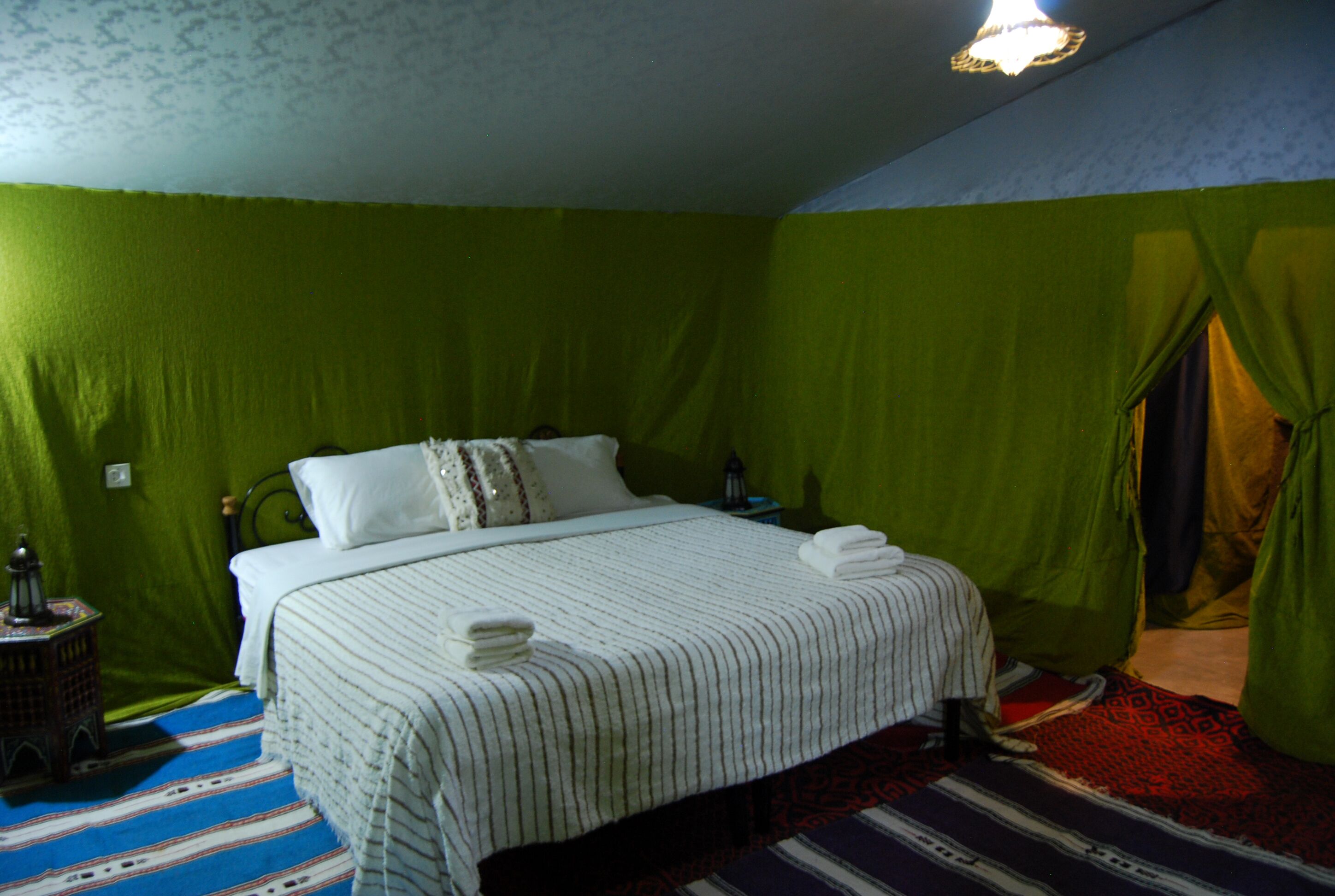 A room with a bed, green walls, a patterned rug, and a small table with a lamp.