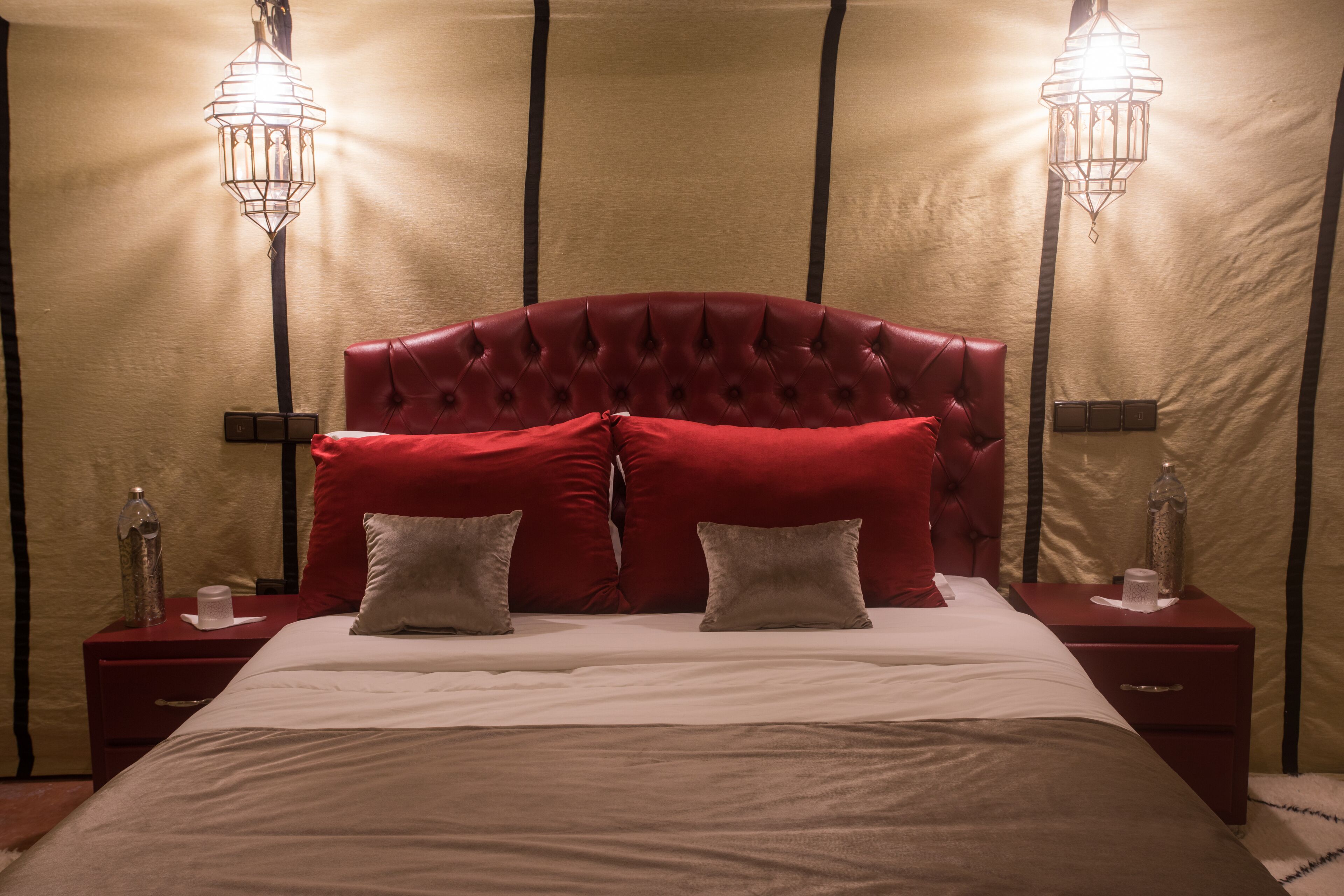 Een slaapkamer met een gestoffeerd hoofdeinde, rode en witte kussens, twee nachtkastjes met lampen en een beige muur.