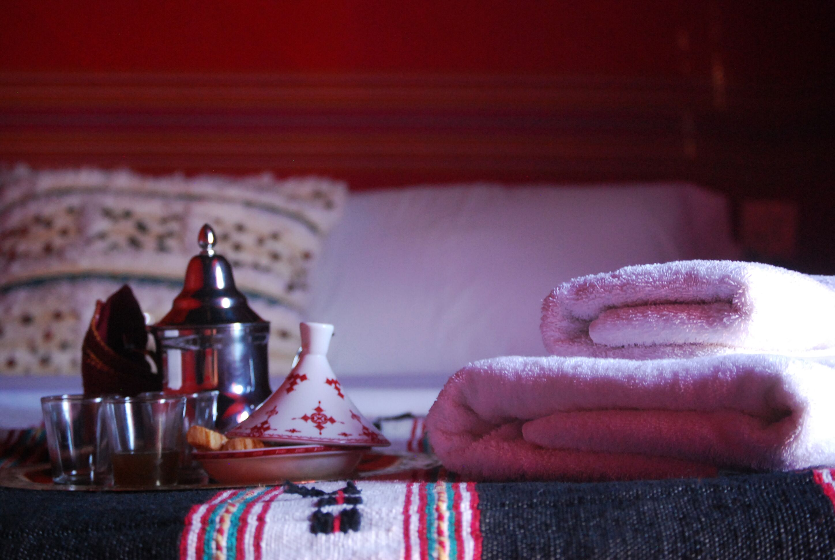 A neatly arranged breakfast setting with a traditional Moroccan tagine, a teapot, glasses, and a plate of pastries on a bed with a striped blanket.