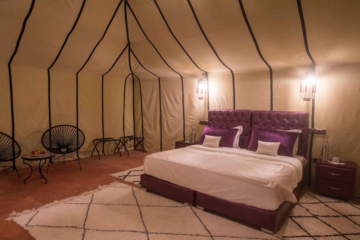 Quarto estilo tenda com uma cama, duas cadeiras, uma mesinha e um criado-mudo.