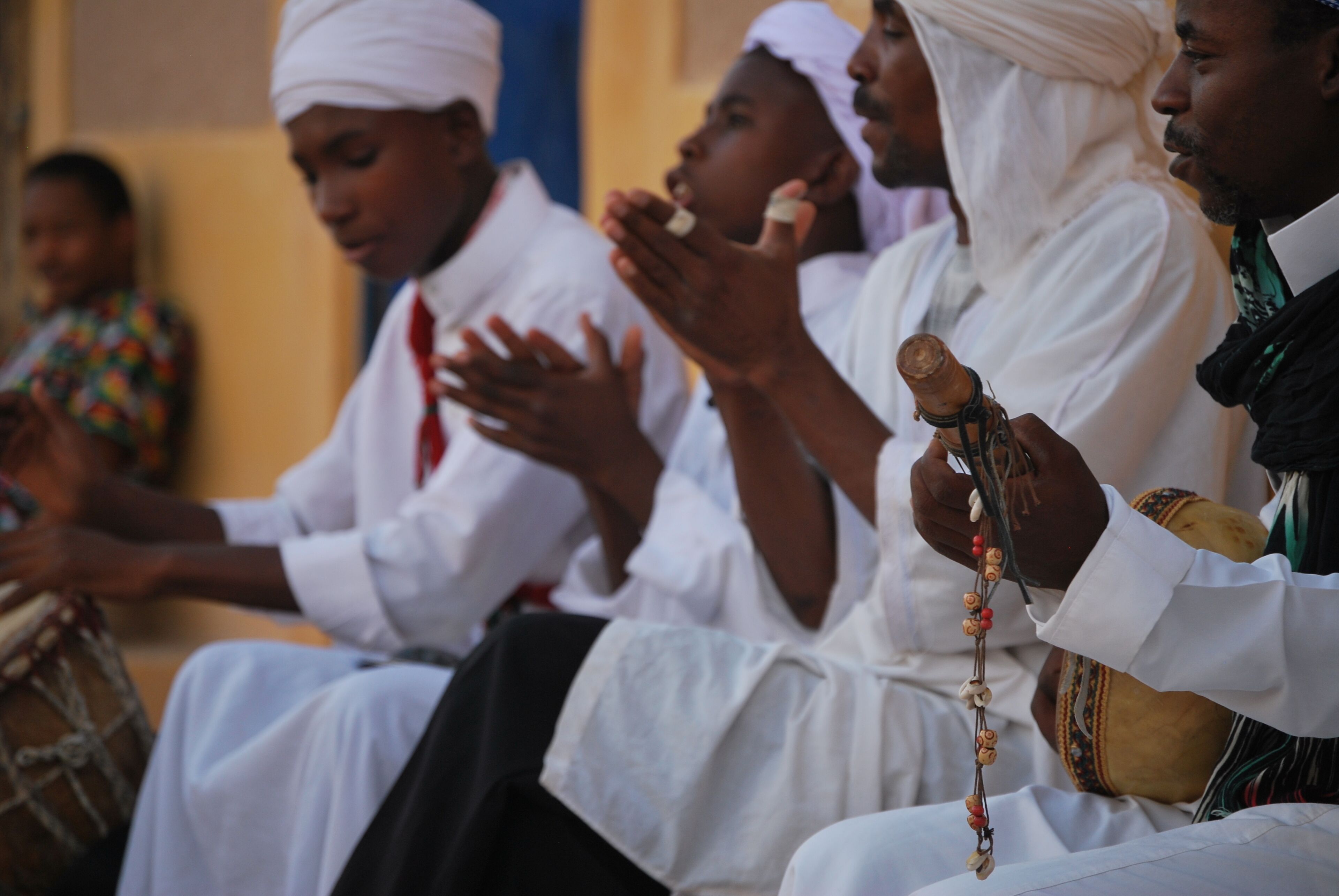 Un groupe de personnes vêtues de tenues traditionnelles blanches, dont certaines tiennent des instruments de musique, sont assises en rangée.