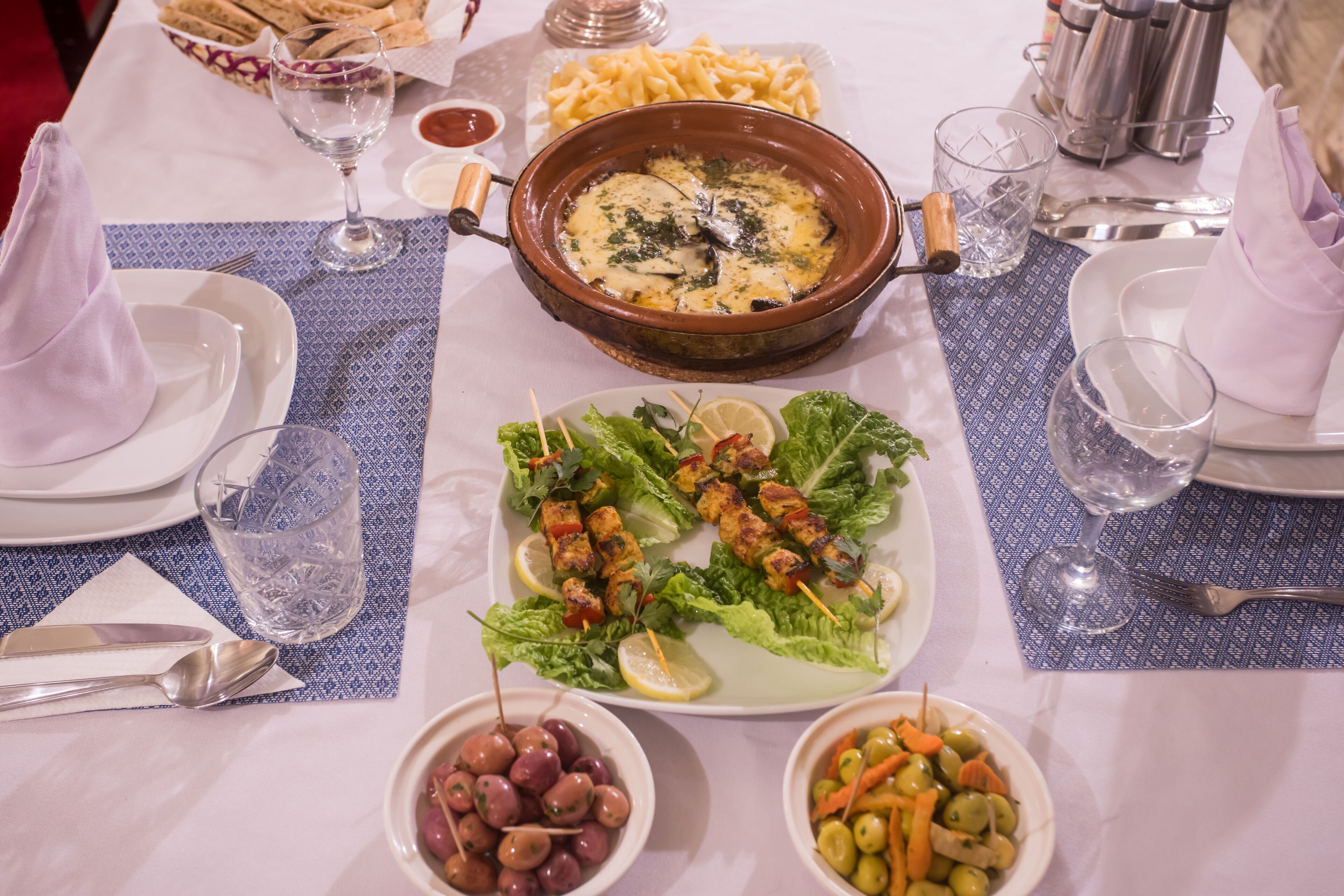 A table set with plates, glasses, and a variety of dishes including grilled skewers, olives, and a bowl of pasta.