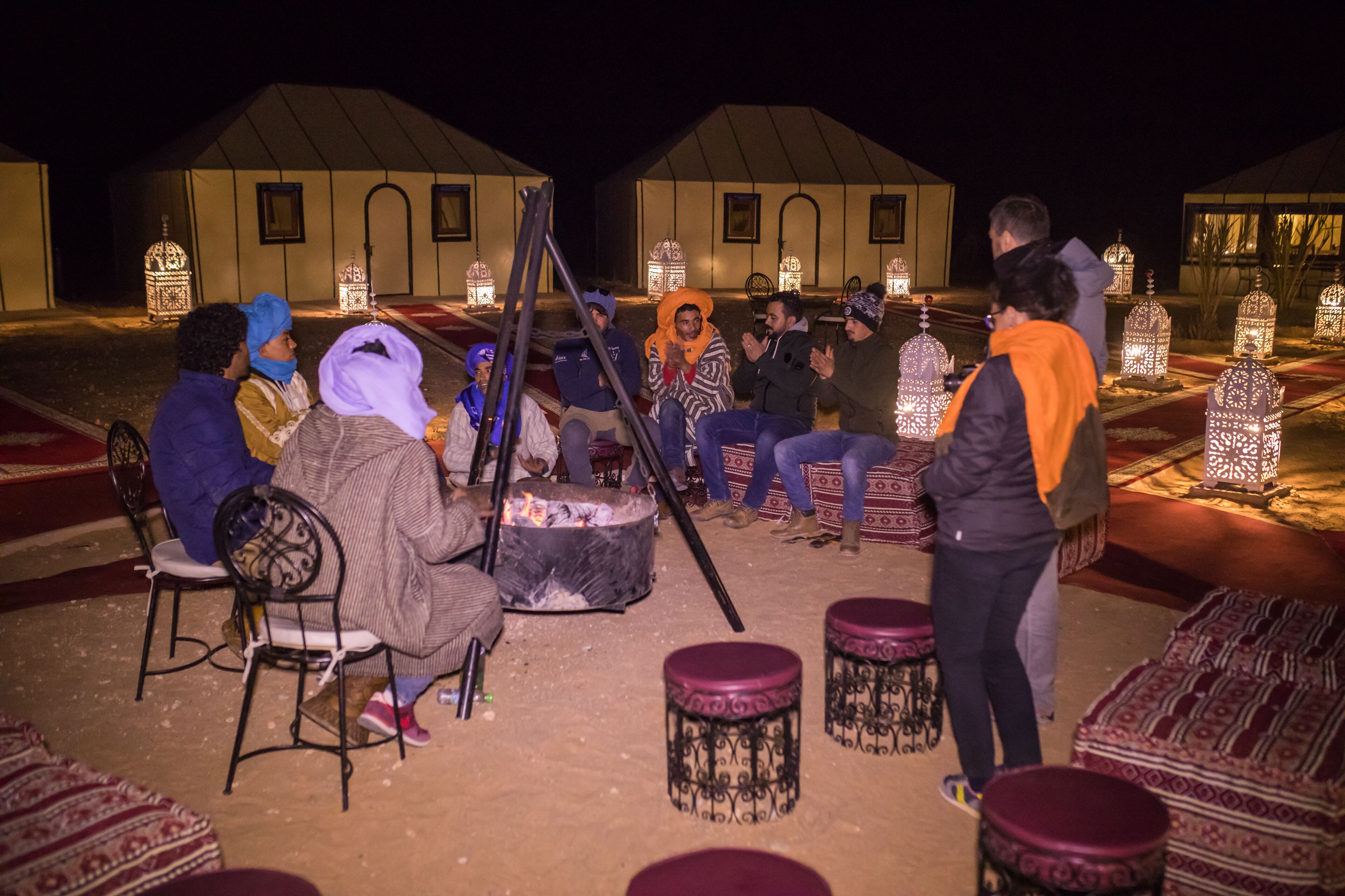 A group of people seated around a fire pit in an outdoor setting with tents and decorative lanterns in the background.