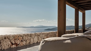 Un mur de pierre offrant une vue sur la mer et les montagnes. Des coussins de couleur beige sont posés sur une surface sombre.