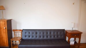 A room with a dark-colored sofa, a wooden chair, a small wooden table with a lamp, and a cabinet. - Incredible Location One Bedroom Apartment (Potts Point)