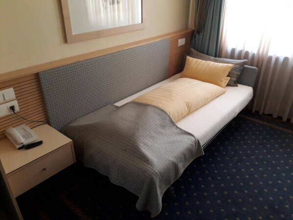 A hotel room with a bed, a bedside table with a phone, and a window with curtains.