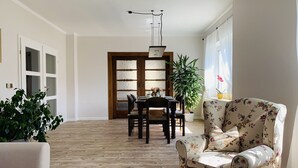 Un salon spacieux doté d’un parquet en bois, un coin repas avec une table et des chaises en bois, un fauteuil à motifs floraux et une plante en pot.