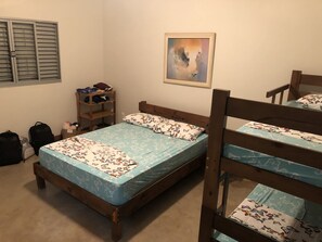 A single bed with a floral bedspread, a wooden headboard, a small shelf with clothes, a framed picture on the wall, and a window with shutters. - Casa Relax Brotas (Brotas)