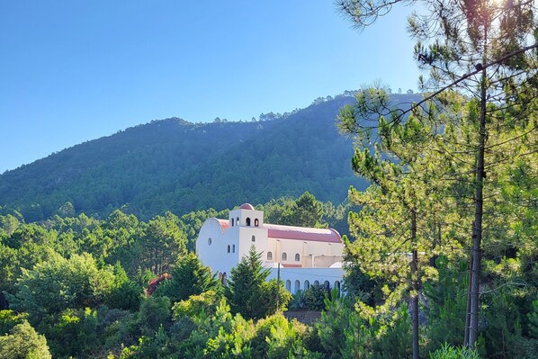 A white building with a red roof surrounded by dense green trees and a hill in the background.