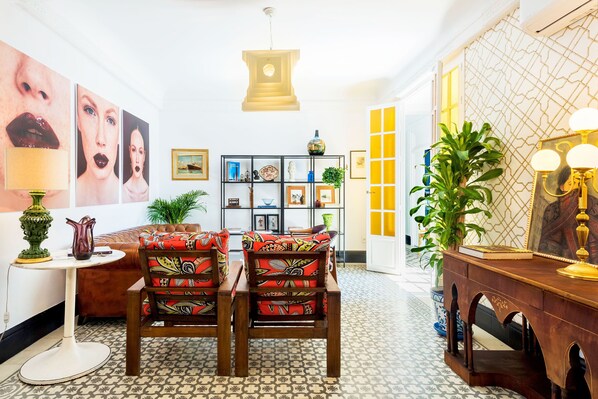 Une pièce avec un sol à motifs, un canapé, deux fauteuils, une petite table, une bibliothèque et un miroir agrémenté d’appliques murales.