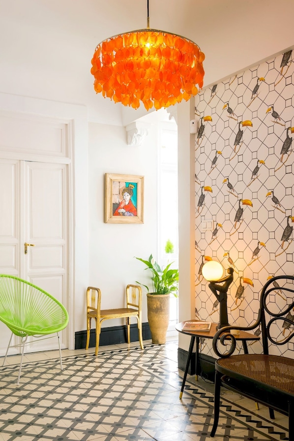 A room with a patterned floor, a green chair, a wooden chair, a table with a lamp, and a wall with a toucan design. - Genteel Home Romanilla (Granada)