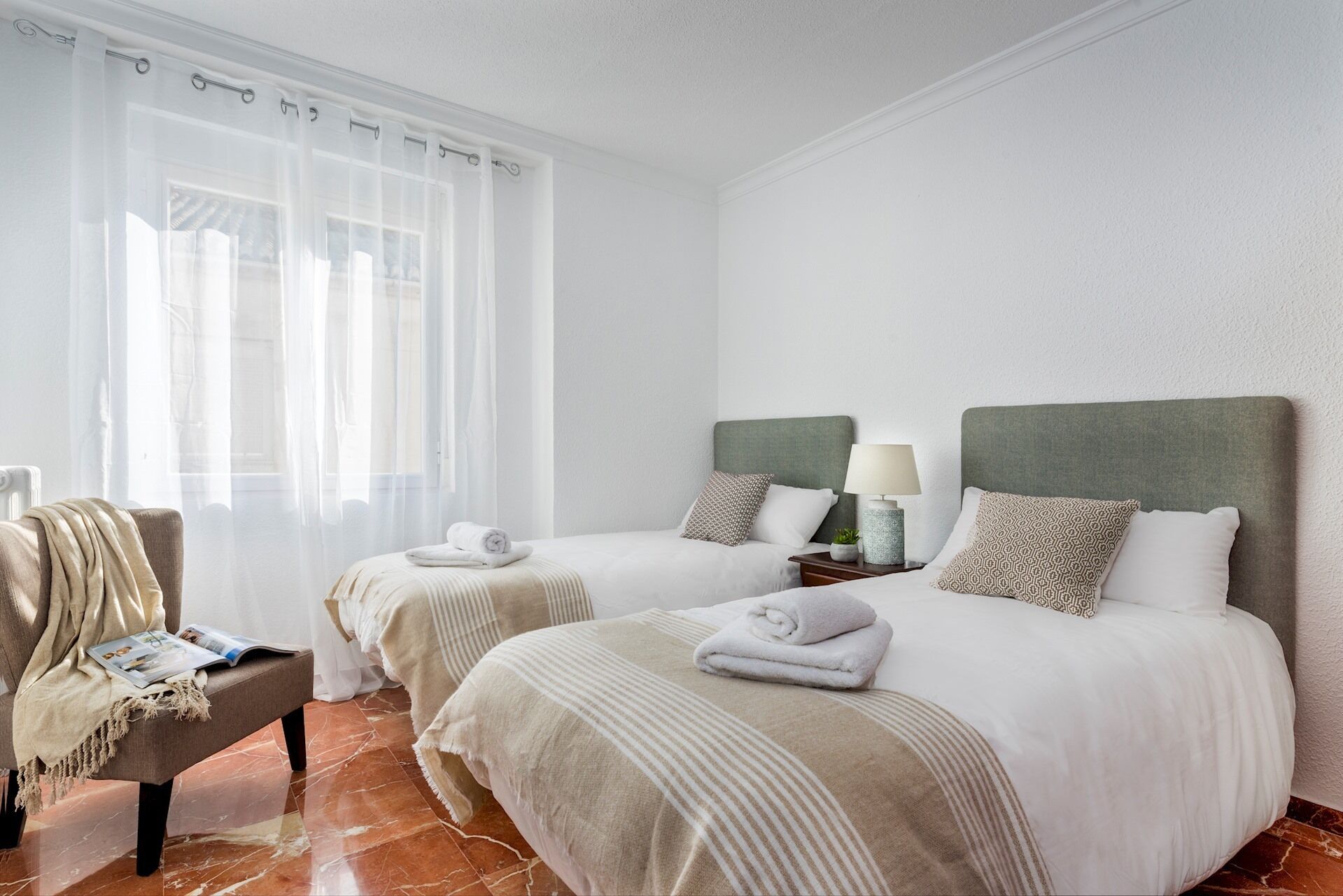 A hotel room with two beds, a chair, a table with a lamp, and a window with sheer curtains.