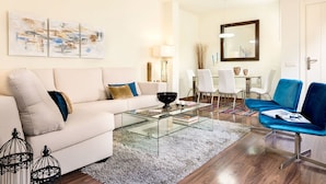 A modern living room with a white sofa, a glass coffee table, a blue chair, and a dining area with white chairs and a table. - Genteel Home Francos Terrace V (Seville)