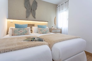 Un dormitorio con dos camas, una gran pieza decorativa de plumas en la pared, una mesita de noche con lámpara y una ventana con cortinas.