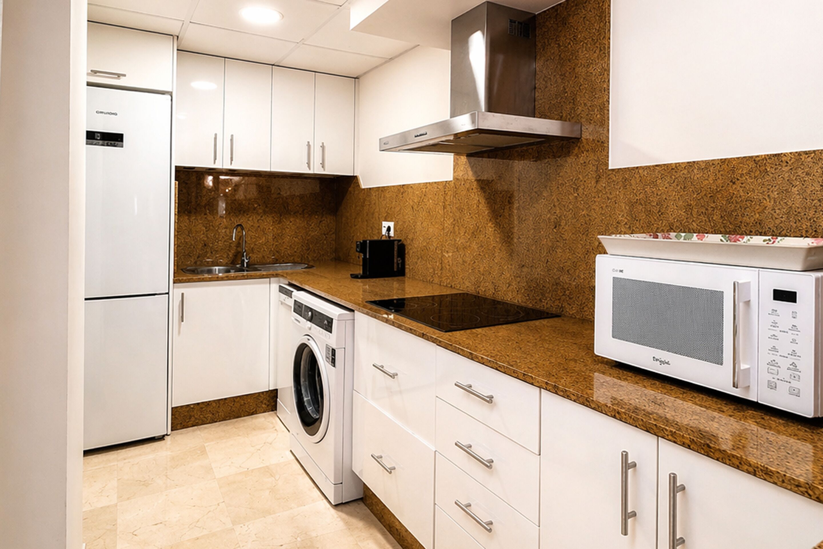 A compact kitchen with white cabinets, a built-in washing machine, a microwave, and a stainless steel range hood.
