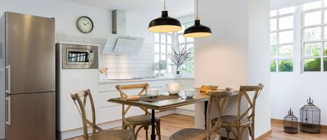 A modern kitchen with a stainless steel refrigerator, a microwave, and a wooden dining table with chairs. There are pendant lights above the table and a window with a view of greenery.