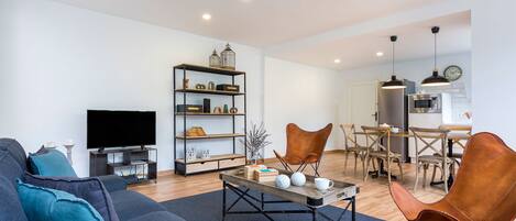 A modern living room with a sofa, coffee table, and a TV. There is a dining area with chairs and a table.