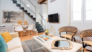 A modern living room with a staircase, a flat-screen TV, and a dining area with wicker chairs.