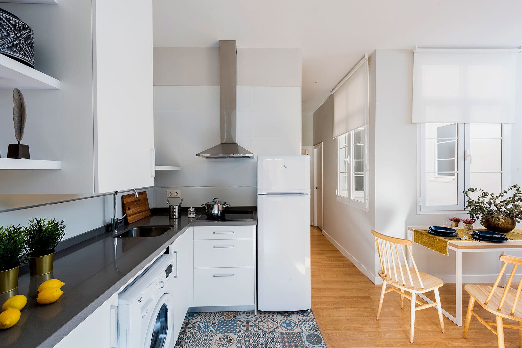 A compact kitchen with white cabinetry, a stainless steel range hood, a built-in oven, and a refrigerator. There is a wooden dining table with chairs, a small rug, and a window with white shutters.