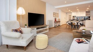 Una sala de estar moderna con un televisor de pantalla plana, un sofá, una mesa de centro y una zona de comedor con sillas y una mesa pequeña.