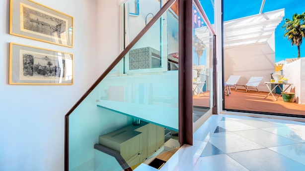 Ein modernes Interieur mit einer Glastreppe, gerahmten Bildern an der Wand und Blick auf die Außenterrasse mit Liegestühlen.