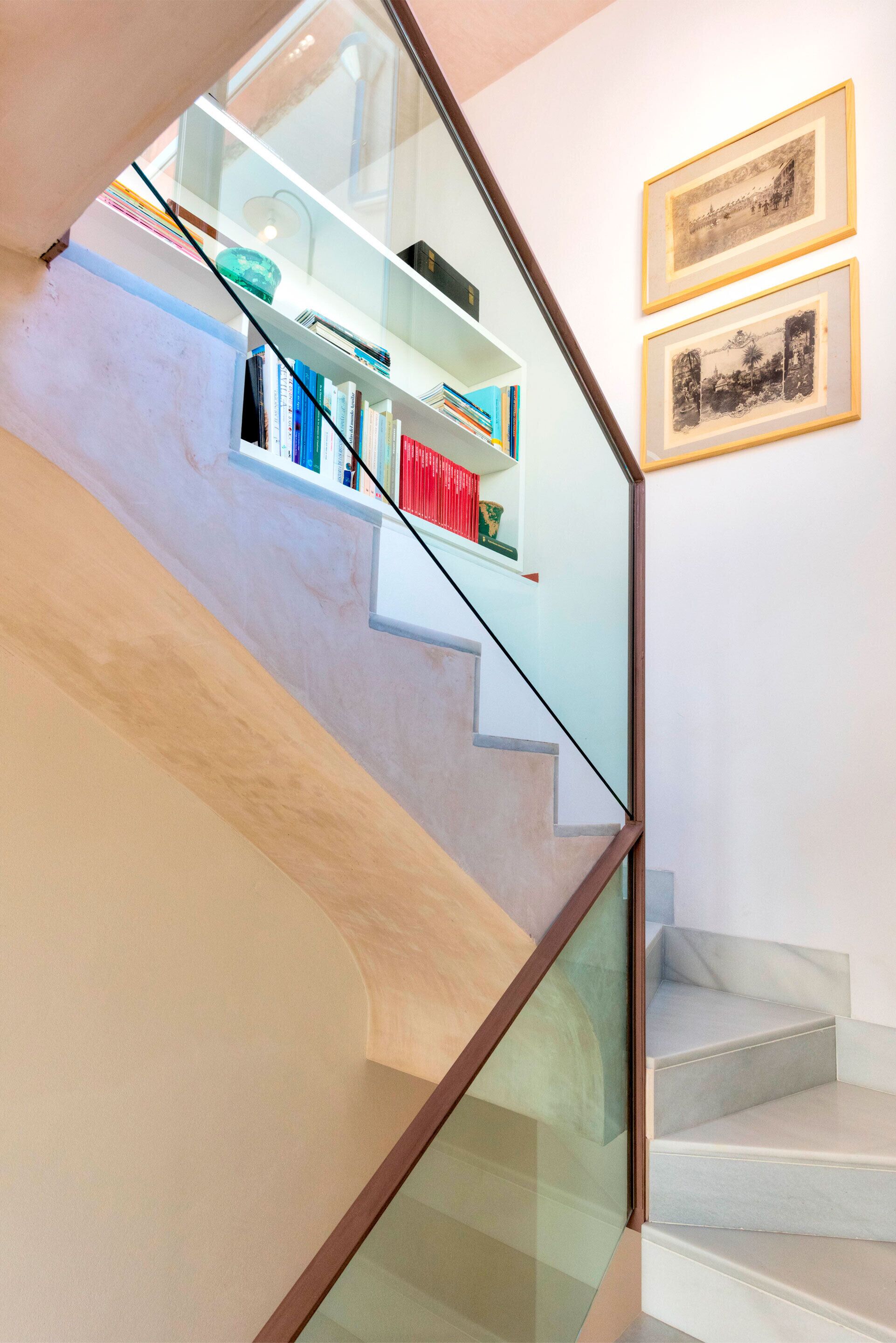 Un escalier avec une rampe en verre, une bibliothèque et deux tableaux encadrés accrochés au mur.