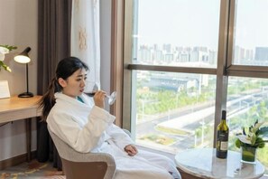 A person in a white robe sitting by a window with a city view, a table with a bottle, and a lamp on a desk. - Montriche International Hotel (WEIFANG)