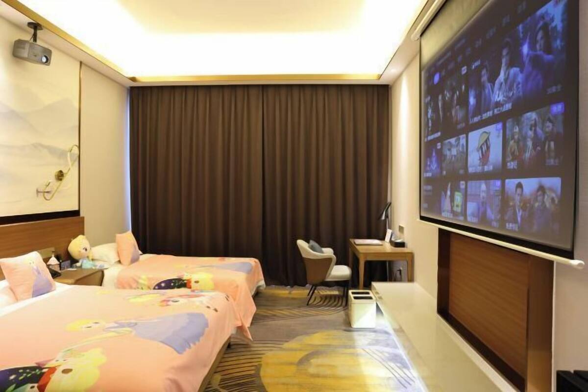 Une chambre d’hôtel avec deux lits, un bureau, une chaise, un écran de projection et une grande fenêtre avec des rideaux.