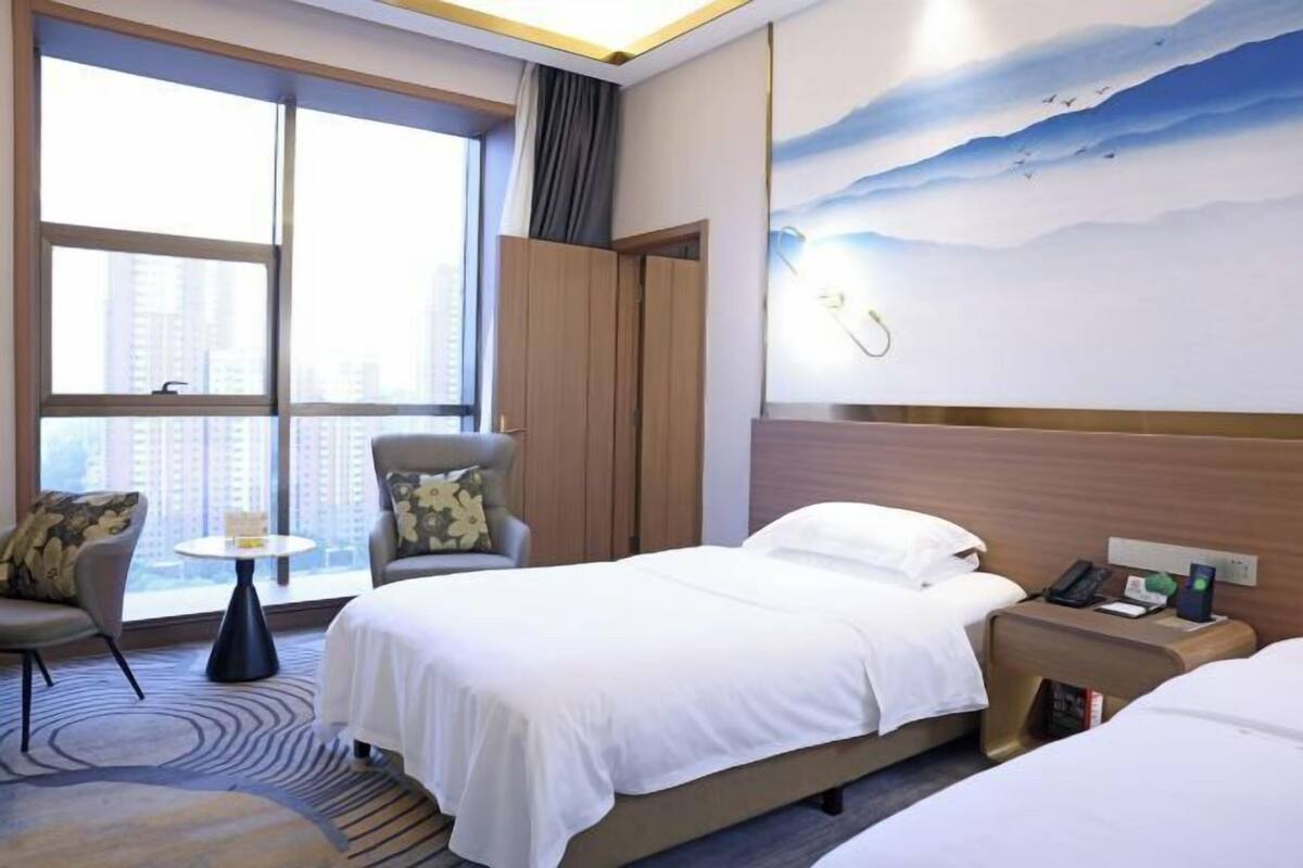 Une chambre d’hôtel avec deux lits, une petite table, une chaise, une armoire et une grande fenêtre donnant sur des immeubles.