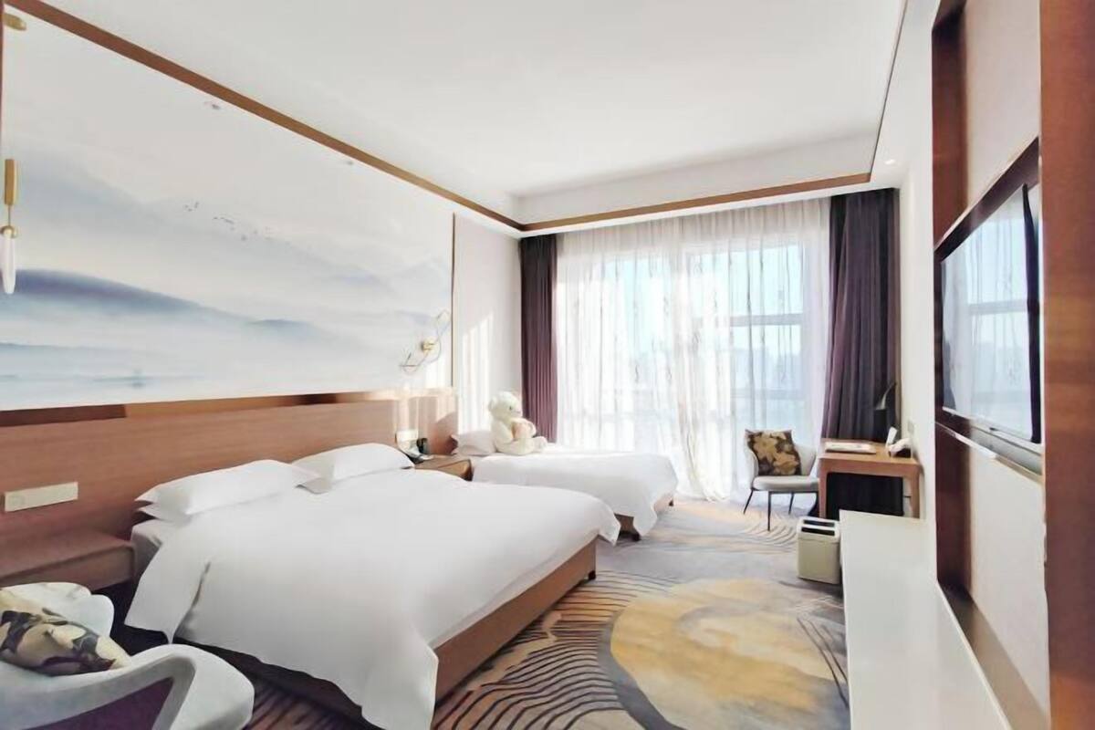 Une chambre d’hôtel avec deux lits, un bureau, une chaise, une grande fenêtre avec des rideaux et un téléviseur fixé au mur.