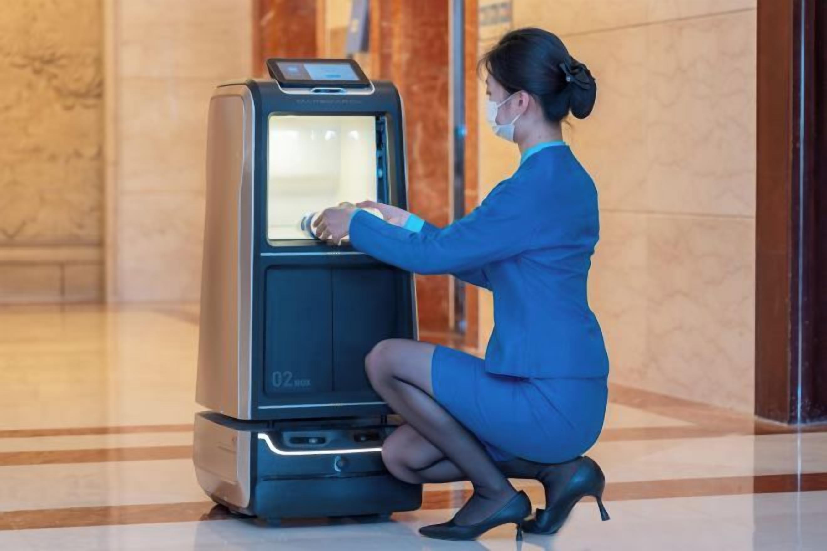 A woman in a blue dress using a self-service kiosk in a lobby with marble flooring and a glass door in the background.