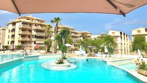 A swimming pool with a central island, surrounded by lounge chairs and umbrellas, with a backdrop of multi-story buildings. - *****Fewo Mariposa - luxury beach penthouse 200sqm, great sea view, free garage (Guardamar)