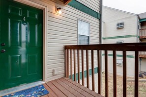 Une porte verte avec un paillasson, une terrasse en bois et un mur extérieur blanc agrémenté d’une fenêtre.