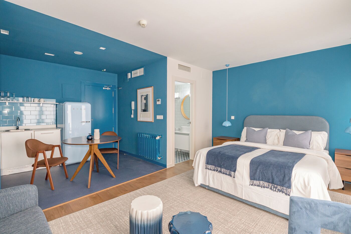 A modern hotel room with a blue color scheme, a bed with a grey headboard, a small dining area with a table and chairs, and a kitchenette with a fridge and a sink.