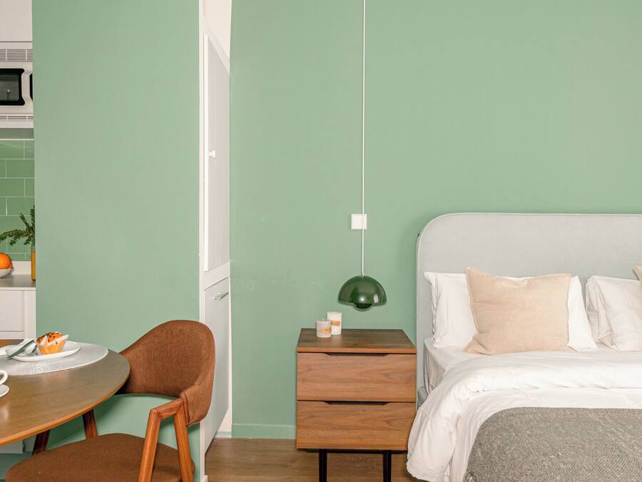 A room with a bed, a desk with a chair, a small table with a bowl of fruit, and a green pendant light.