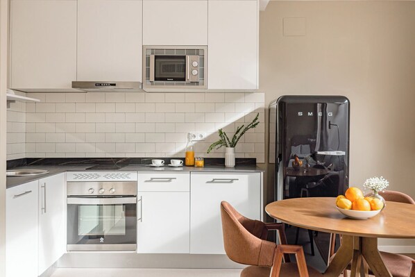 Una cocina moderna con armarios blancos, microondas integrado, horno de acero inoxidable y refrigerador negro. Hay una mesa de comedor con un cuenco de fruta y una planta en maceta sobre la encimera.