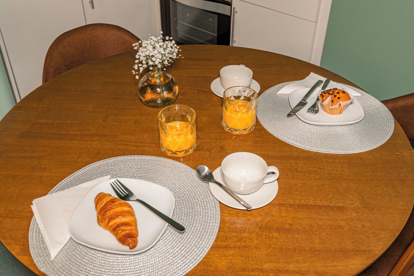 A breakfast setting with croissants, orange juice, and a small vase of flowers on a wooden table.