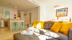 A modern living room with a grey sofa, yellow cushions, and a coffee table with a tray. A kitchen with a dishwasher and bar stools is visible in the background. - Sebastian Patio by Hello Homes Sitges (Sitges)