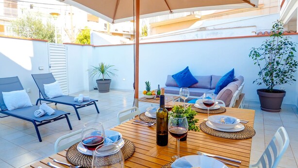 A sunlit patio with a wooden table set for dining, lounge chairs, and a sofa with cushions. - Sebastian Patio by Hello Homes Sitges (Sitges)