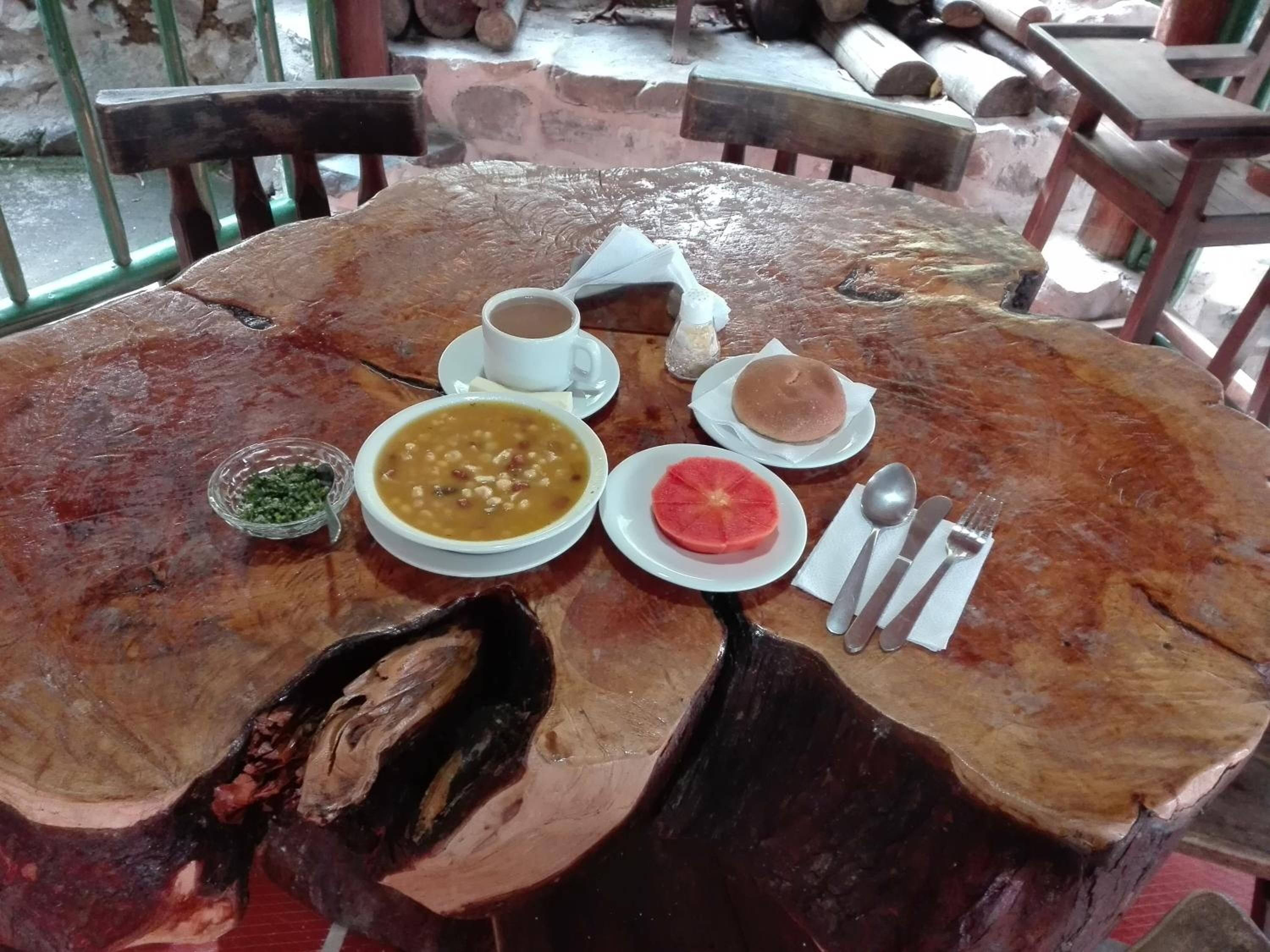Une table en bois rustique, dressée à l’extérieur, avec un repas composé d’une soupe, de pain et d’une tranche de fruit, le tout agrémenté d’une vue sur des troncs d’arbres et une rambarde en métal.