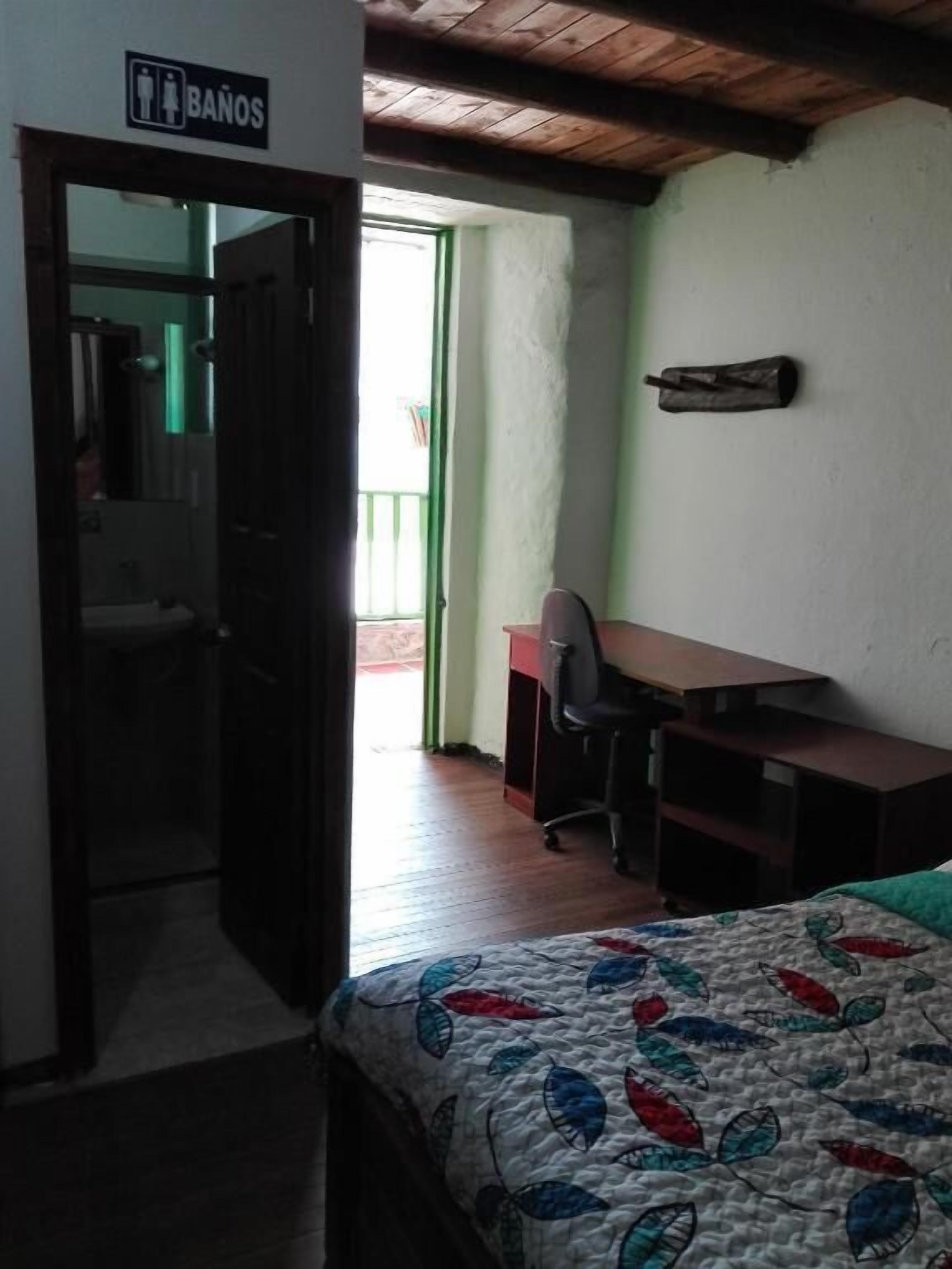 Habitación con cama, escritorio y silla. Se ve un cartel que indica el baño.