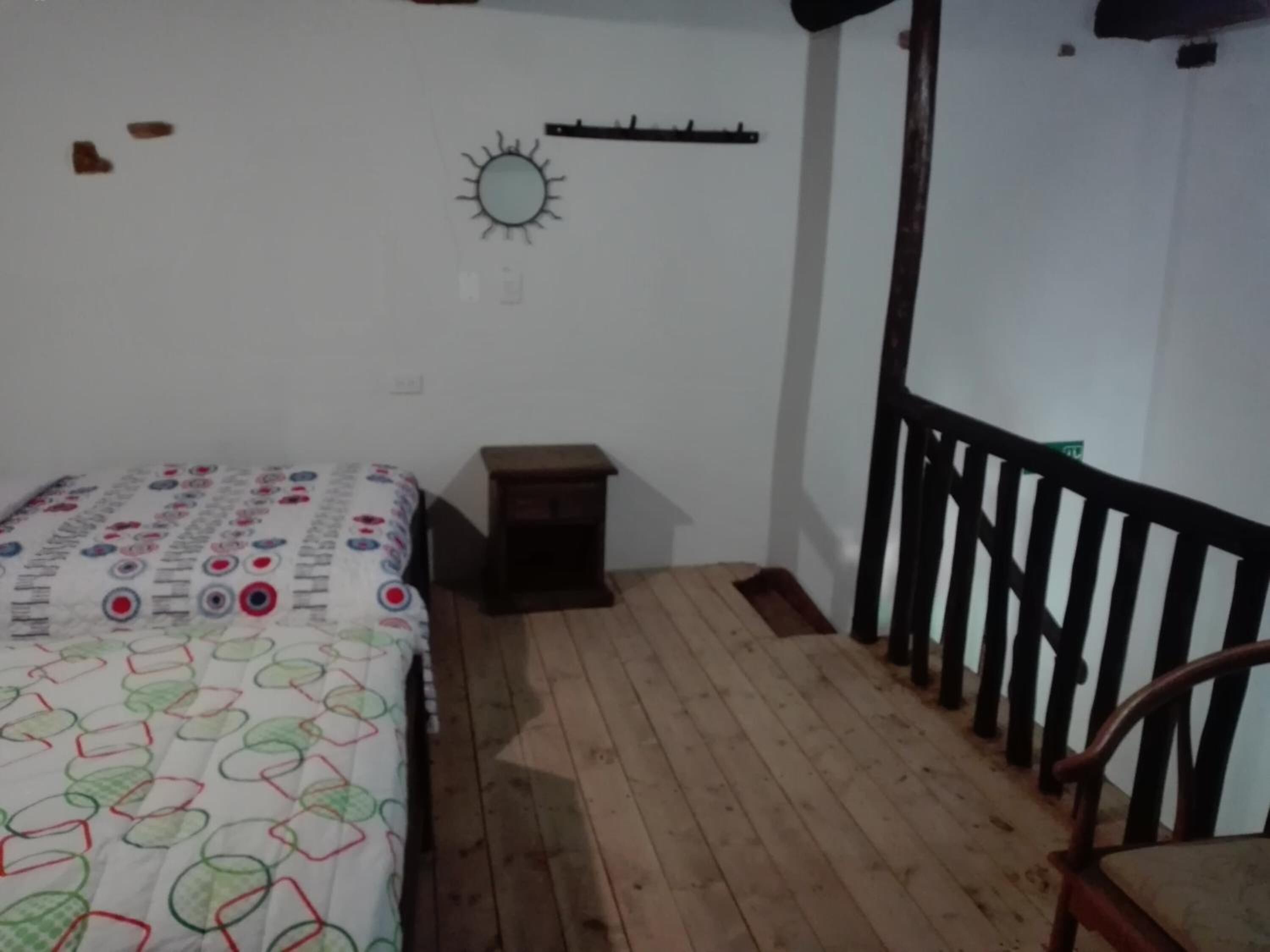 A bedroom with a bed, a small wooden stool, a wooden floor, a white wall with a sun decoration, and a black railing.