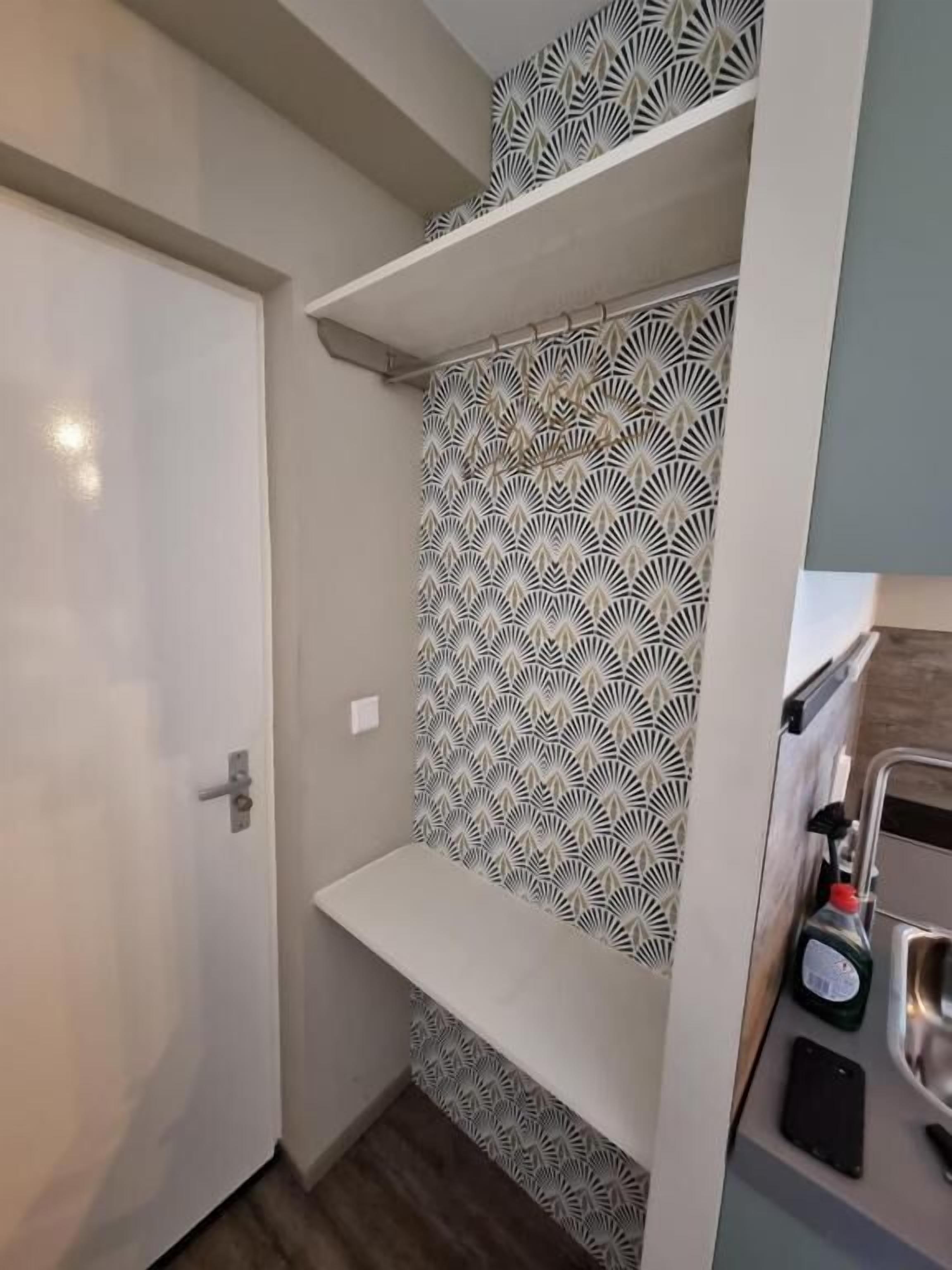 Une salle de bain avec des carreaux décorés, une porte blanche et une étagère sur laquelle se trouvent une bouteille et un téléphone intelligent.