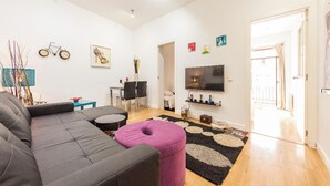Un salon moderne avec un canapé gris, un pouf rose, un téléviseur et un coin repas comprenant des chaises et une table.